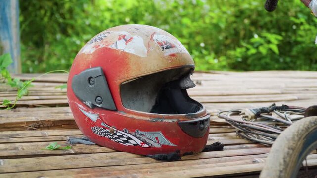 Close up of weathered red motorcycle helmet with racing decals resting on wooden planks. scene includes tangled cables and green foliage in background suggesting outdoor setting with natural