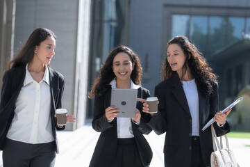Three female colleagues in matching black business suits, talking as they walk in step, move as a group across a sunlit forecourt carrying a tablet, coffee cups and documents. © ultramansk