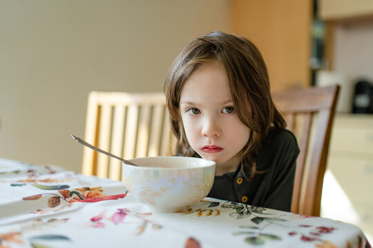 Young child sitting at a dining table and looking reluctant to eat his meal. Concept of picky eating, poor appetite and difficulties feeding children at home.