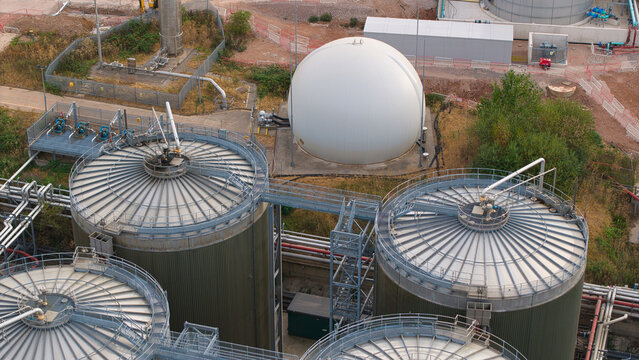 Aerial view of the wastewater treatment plant featuring large cylindrical storage tanks and a white spherical gas holder surrounded by industrial infrastructure in Wanlip, England, United Kingdom.