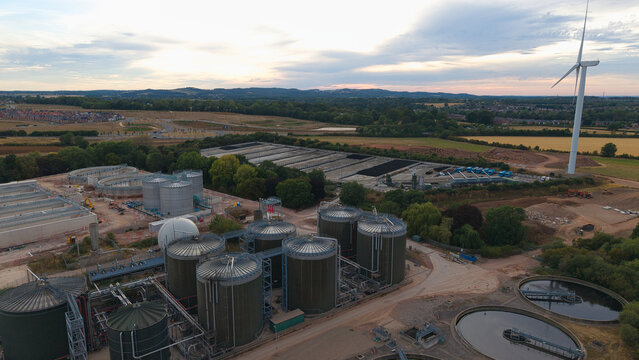 Aerial view of a wastewater treatment plant featuring large industrial tanks, circular sedimentation basins, and a wind turbine in Wanlip, England, United Kingdom.