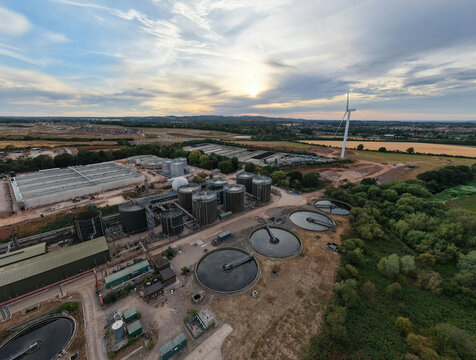 Aerial view of the wastewater treatment plant featuring large circular sedimentation tanks, industrial infrastructure, and a wind turbine in Wanlip, England, United Kingdom.