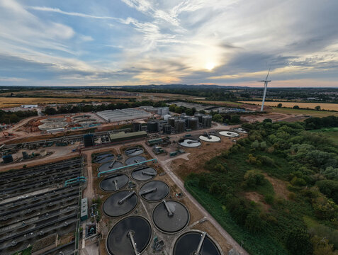 Aerial view of the wastewater treatment plant featuring circular sedimentation tanks, industrial infrastructure, and a wind turbine under a cloudy sky in Wanlip, England, United Kingdom.