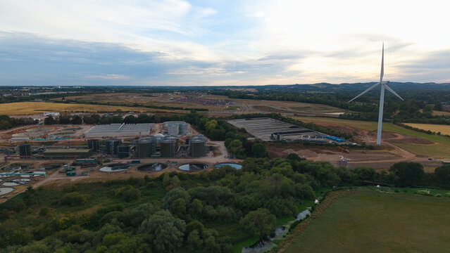 Aerial view of the wastewater treatment plant featuring large industrial tanks, settling ponds, and a wind turbine set against a rural landscape in Wanlip, England, United Kingdom.