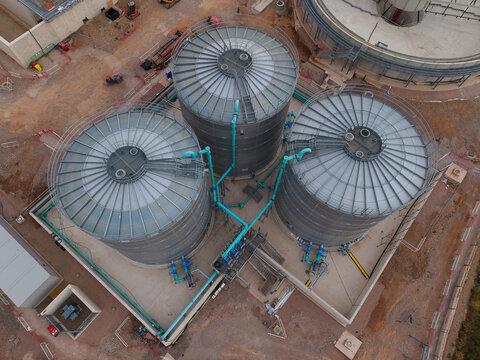 Aerial view of the wastewater treatment plant featuring three large circular metal tanks connected by blue piping in an industrial facility in Wanlip, England, United Kingdom.