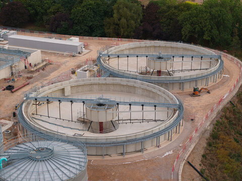 Aerial view of wastewater treatment plant featuring large circular concrete sedimentation tanks with mechanical bridges and surrounding industrial infrastructure in Wanlip, England, United Kingdom.