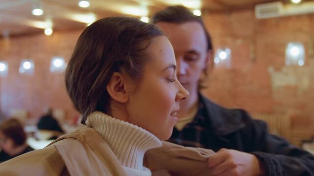 Close up of woman wearing white turtleneck and man helping her put on beige coat in cozy cafe with brick walls warm lighting tables chairs other patrons. Woman adjusts hair while interacting