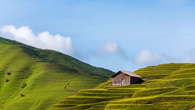 Static aerial tracking shot of wooden hut nestled on layered terraced green hills overlooking rural mountainside under blue sky with scattered clouds
