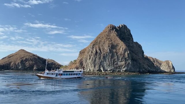 Traditional wooden phinisi boat sailing on blue sea waters towing a small dinghy near scenic arid hills at Komodo National Park, Indonesia.