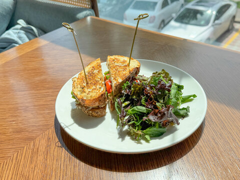 Golden Toasted Sandwich Halves, Served With Fresh Mixed Leaf Salad On White Plate, Rest On Wooden Table Beside Bright Window Inside Modern Cafe, With Soft Sunlight And Blurred Street