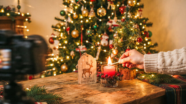 Una mano enciende con una cerilla una vela sobre una mesa de madera decorada, junto a un peque&ntilde;o pino navide&ntilde;o, casita de jengibre y un &aacute;rbol iluminado de fondo.