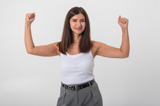 Smiling woman standing against a white background, raising her arms and flexing her biceps, conveying feelings of empowerment, determination, and success