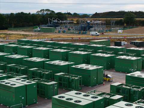 Aerial view of a large battery energy storage system facility featuring rows of green containers and an electrical substation in the background in Birmingham, England, United Kingdom.