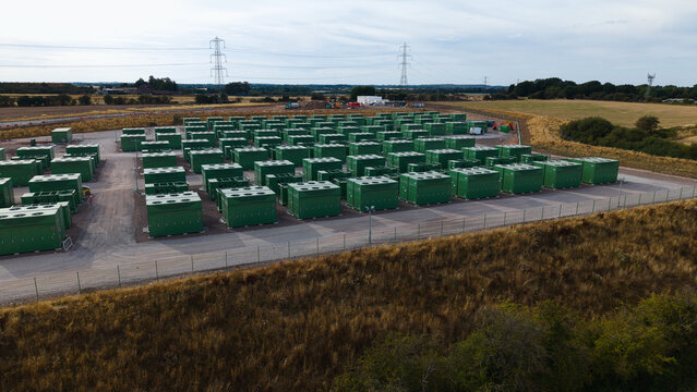 Aerial view of a large-scale battery energy storage system facility with rows of green container units, gravel ground, and surrounding fields in Birmingham, England, United Kingdom.