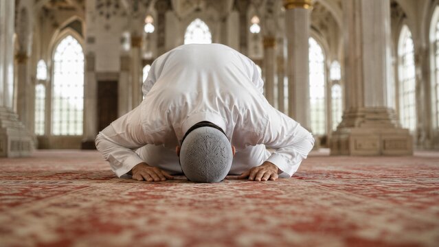 Muslim man in white clothing performs prostration during prayer inside a grand mosque with large windows and ornate architecture.