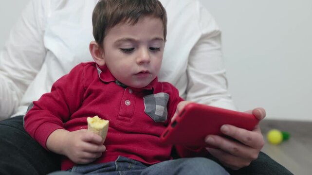 Little boy watching smartphone and eating banana on mother's lap