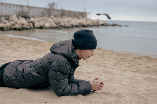 A teenager lies playfully on the sandy autumn beach, fooling around near the calm sea on a cold overcast day by the shore
