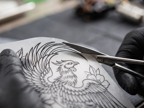 Tattoo Artist Preparing Stencil by Cutting Bird Design with Scissors