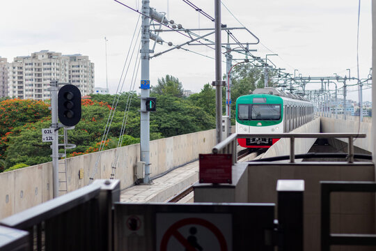 Dhaka, Bangladesh - 26 May 2024: View of the Metro Rail train arriving at the station platform with overhead power lines, signal lights, and urban greenery in the background.