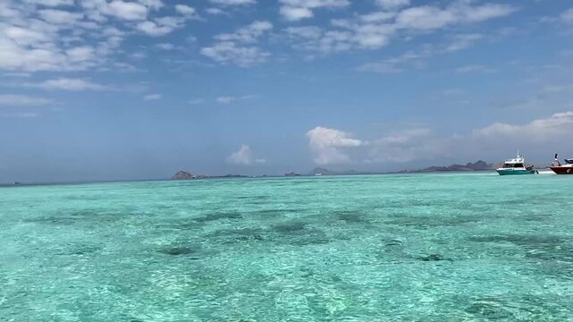 Clear turquoise sea with tourists snorkeling and phinisi boats anchored near islands under a bright blue sky at Taka Makassar Island, Komodo National Park, Indonesia.