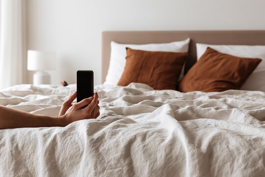 Hand holding phone over unmade bed, soft pillows and white sheets.
