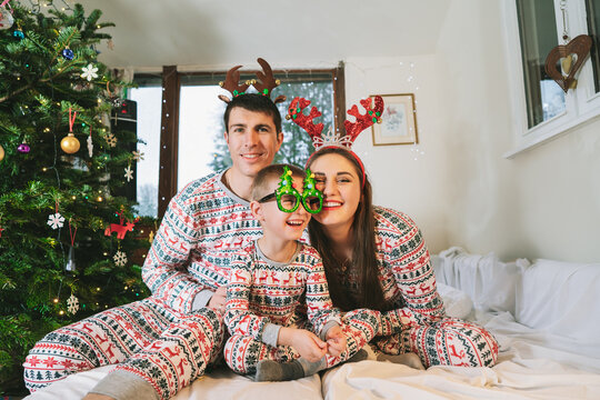 Happy family in pajamas and funny carnaval hats and glasses on bed on Xmas Eve in decorated apartment. Dad, mom, son lying on bed and having fun together. Family celebrating winter holidays at home.