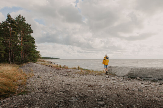 View of a person wearing a yellow jacket standing on a rocky beach by the sea under a cloudy sky in Saxby, Laane maakond, Estonia.