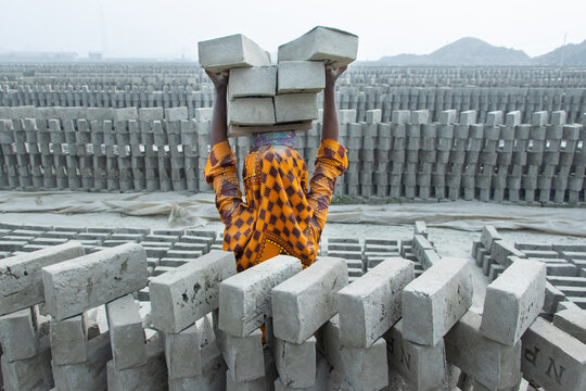 Dhaka, Bangladesh - 24 January 2021: View of a labourer carrying a stack of bricks on her head in a brick field, with rows of drying bricks in the background.