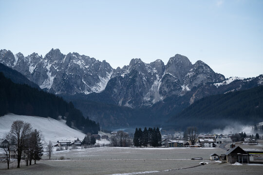 Wide shot of snow covered Gosaukamm mountain range in Gosau valley, Austria. Alpine village surrounded by pine forest and frosted fields in winter.