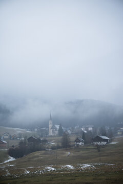 Vertical shot of foggy alpine village with tall church spire surrounded by mist covered hills and light snow in winter. Atmospheric winter landscape in Gosau, Austria.