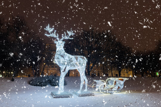 Glowing LED reindeer sculpture and sleigh in park during falling snow. Magical Christmas light decoration on snowy winter night, outdoor Christmas light.