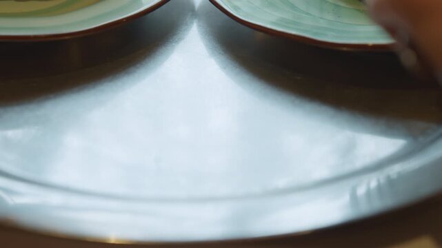 Close up of hand reaching for and picking up silverware from empty plate on table. fork is moved across metallic surface revealing texture details under soft lighting. Plates are visible