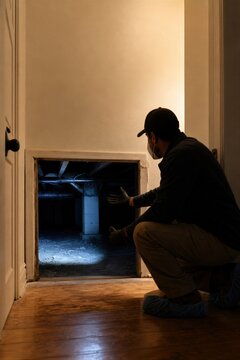 Pest control technician examining crawlspace under residential house. Man inspecting home foundation for termite damage or mold growth.