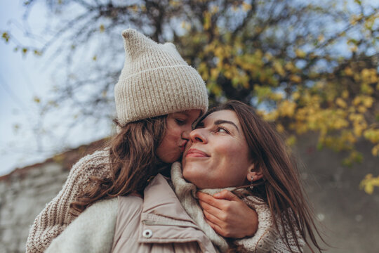 Close-up outdoor portrait of young woman kissed on cheek by little daughter. Family love, parenthood, maternal affection, bonding, tenderness, childhood, autumn lifestyle, warmth, happiness.