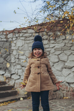 Happy little girl in beige coat and blue hat playing with falling autumn leaves outdoors. Magic of autumn, childhood wonder, happiness, lifestyle.