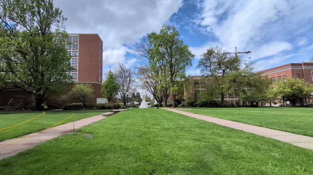 A broad grassy field stretches across a campus, framed by historic buildings, pathways and a construction crane beneath a blue spring sky.