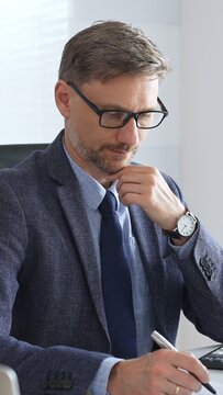 Adult businessman wearing blue suit and tail is concentrating and thinking while working at the desk in a bright, modern office, demonstrating professionalism and efficiency. Business people concept