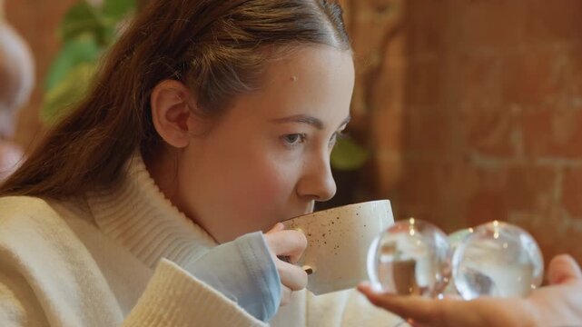 Close up of young woman drinking from cup in indoor setting. Woman interacts with another woman holding clear glass spheres maintaining calm expressions throughout scene. background features