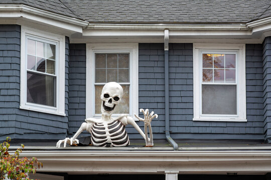 Halloween skeleton decoration emerging from porch roof of gray-blue suburban house