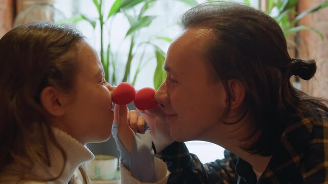 Close up of young girl and man holding red objects near their faces in indoor setting with soft lighting. subjects interact closely maintaining focus on shared activity throughout scene