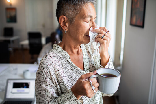 Older woman sneezing into tissue while holding tea at office