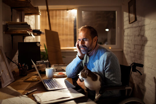 Man in wheelchair working from home office with cat at night