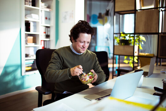 Smiling man eating healthy salad while working on laptop in modern office