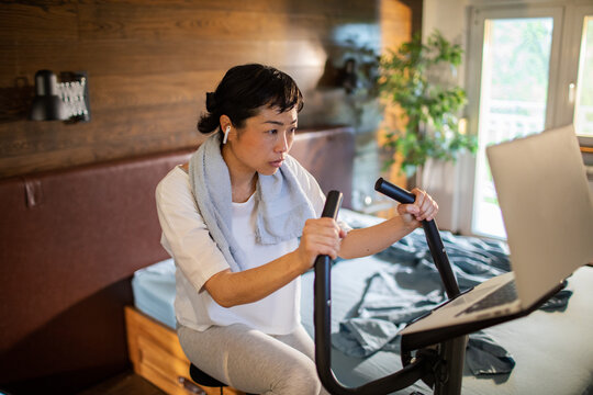Focused woman cycling on exercise bike at home, watching laptop