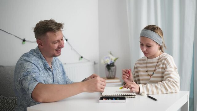 Father and daughter paint with watercolors at their home table. They enjoy a happy creative moment.