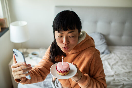 Woman blowing out birthday candle on cupcake during video call in bedroom