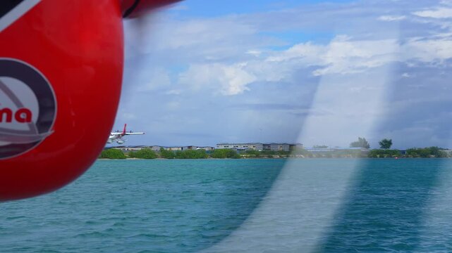 A seaplane lands over turquoise ocean water near an island coastline in the Maldives, viewed from beside another aircraft engine.