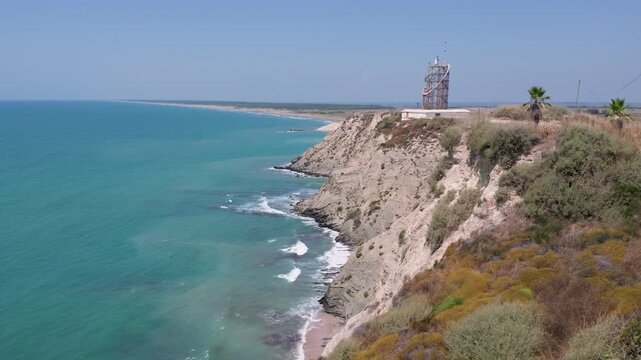 Beautiful view of Karatas Fener Burnu in Adana, Turkey. High rocky cliffs meet the turquoise Mediterranean Sea under a blue sky, featuring a scenic coastal cape landscape with a lighthouse tower.