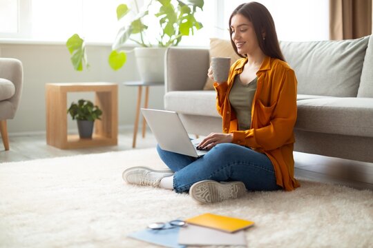 Smiling Young Hispanic Female Working On Laptop While Sitting On Floor With Coffee In Living Room Of Home. Enjoying Relaxed And Comfortable Atmosphere In Modern Interior, Focused On Work.
