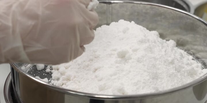 close-up shot of fine white flour or powdered sugar being sifted through a professional stainless steel mesh strainer. The camera captures the detailed texture of the powdery ingredients accumulating 